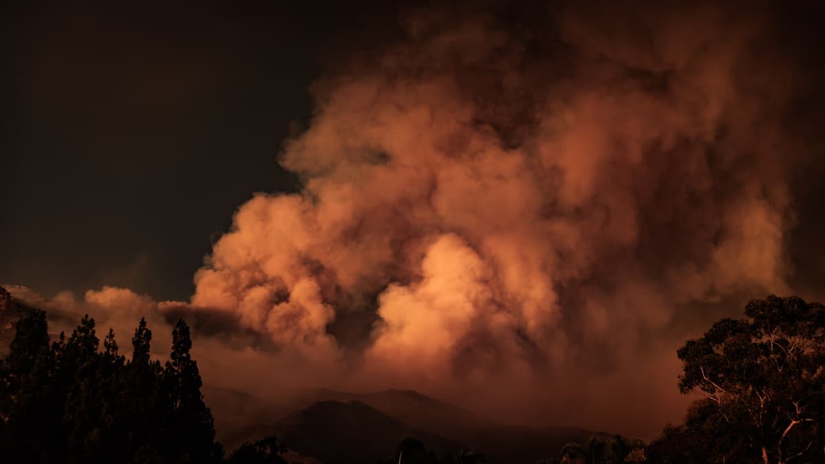 A large plume of smoke rising from a wildfire in a forested mountain landscape